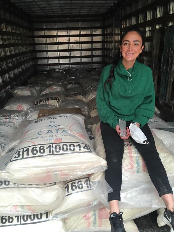 Photograph of Catalina smiling broadly in a green jumper and black jeans sitting on sacks of coffee in a shipping container just before it leaves the port in Cartagena, Colombia